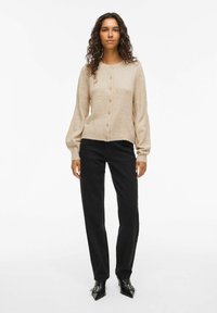 Femme aux cheveux bouclés portant un cardigan beige à boutons, un pantalon noir et des bottines noires à bout pointu, se tenant contre un fond blanc.