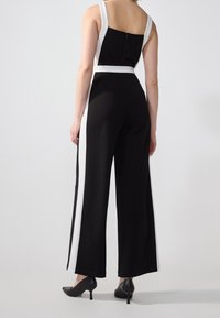 Černý overal s bílými bočními pruhy, širokými nohavicemi a přiléhavým korzetem. Obsahuje zipové zapínání na zadní straně a nastavitelné ramenní popruhy.