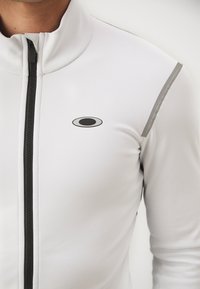 Hvit zip-up jakke med høy krage. Har svart glidelås, grå detaljer på skuldrene og en svart oval logo på brystet. Glatt tekstur.