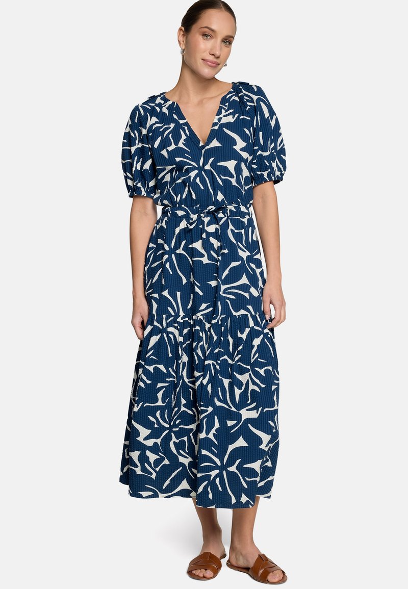 Femme portant une robe midi fleurie bleue et blanche avec manches bouffantes et taille nouée, associée à des sandales slides marron.
