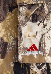 Υφασμα με καμουφλάζ σε γήινες αποχρώσεις του καφέ και του μπεζ. Περιέχει λογότυπο της Adidas σε λευκό και κόκκινο κέντημα. Ανώμαλη επιφάνεια.
