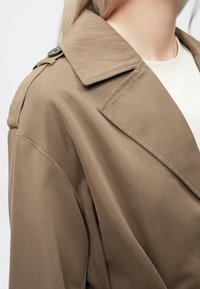 Brown trench coat with a structured collar and shoulder button detail, featuring smooth fabric texture and tailored fit.