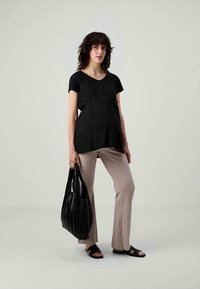 Donna incinta che indossa un top nero e pantaloni beige, con una grande borsa nera, in piedi contro uno sfondo chiaro e semplice.
