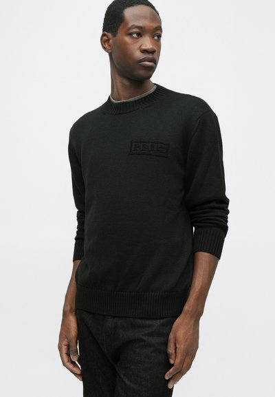 Jaunas vyras juodame megztinyje ir tamsiose kelnėse stovi prieš paprastą šviesią fono, žiūrėdamas šoną.