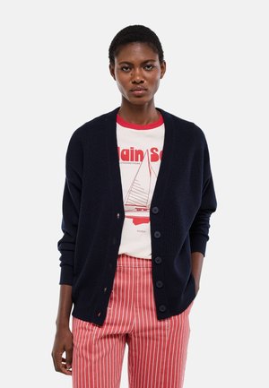 Femme portant un cardigan bleu marine boutonné sur un T-shirt blanc à motif et un pantalon à rayures rouges et blanches, debout devant un fond uni.