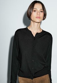 Frau mit kurzen braunen Haaren, die eine schwarze, geknöpfte Strickjacke und braune Hosen trägt, steht vor einem grauen Hintergrund.