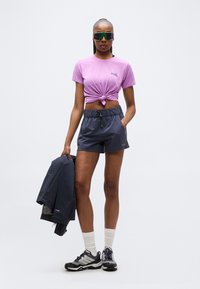 Γυναίκα με μοβ μπλουζάκι δεμένο στον κόμπο, σκούρο μπλε σορτς, λευκές κάλτσες, μαύρα αθλητικά παπούτσια, αντανακλαστικά γυαλιά ηλίου, κρατώντας ένα σκούρο μπλε τζάκετ στο δεξί χέρι.