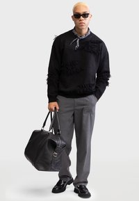 Black sweater with fringe detailing, gray trousers, black leather duffel bag, and black shoes. Accessories include sunglasses and a patterned scarf.