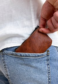 Hnědá kožená kapsička na zip je zasouvána do zadní kapsy světle modrých džínsů. Texturovaný povrch, obdélníkový tvar, viditelný zip.