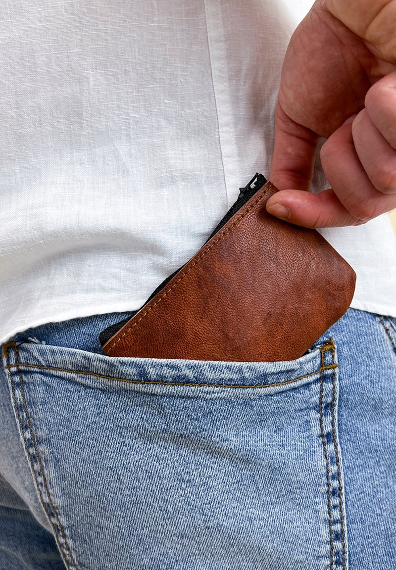 Hnědá kožená kapsička na zip je zasouvána do zadní kapsy světle modrých džínsů. Texturovaný povrch, obdélníkový tvar, viditelný zip.