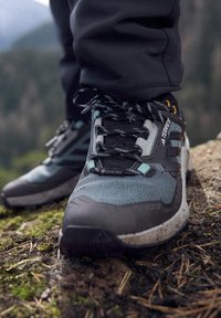Gros plan sur une personne portant des chaussures de randonnée Gore-Tex noires et grises, debout sur un sol moussu en plein air.