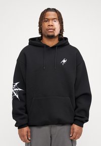 Felpa nera con cappuccio e tasca frontale, impreziosita da un logo di ragno bianco sul petto e un design a ragnatela sulla manica sinistra. Materiale in cotone, vestibilità comoda.