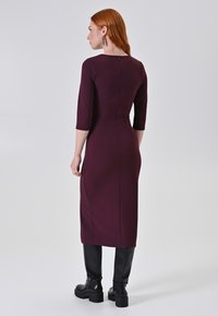 Marlowe jurk in diep bordeauxrood, aansluitende vorm, knielang, met driekwartmouwen en een discrete ritssluiting aan de achterkant. Zwarte knielange laarzen.