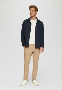 Marineblauw overhemd met knopen over een witte shirt, gecombineerd met beige broek en witte sneakers, staande tegen een neutrale achtergrond.