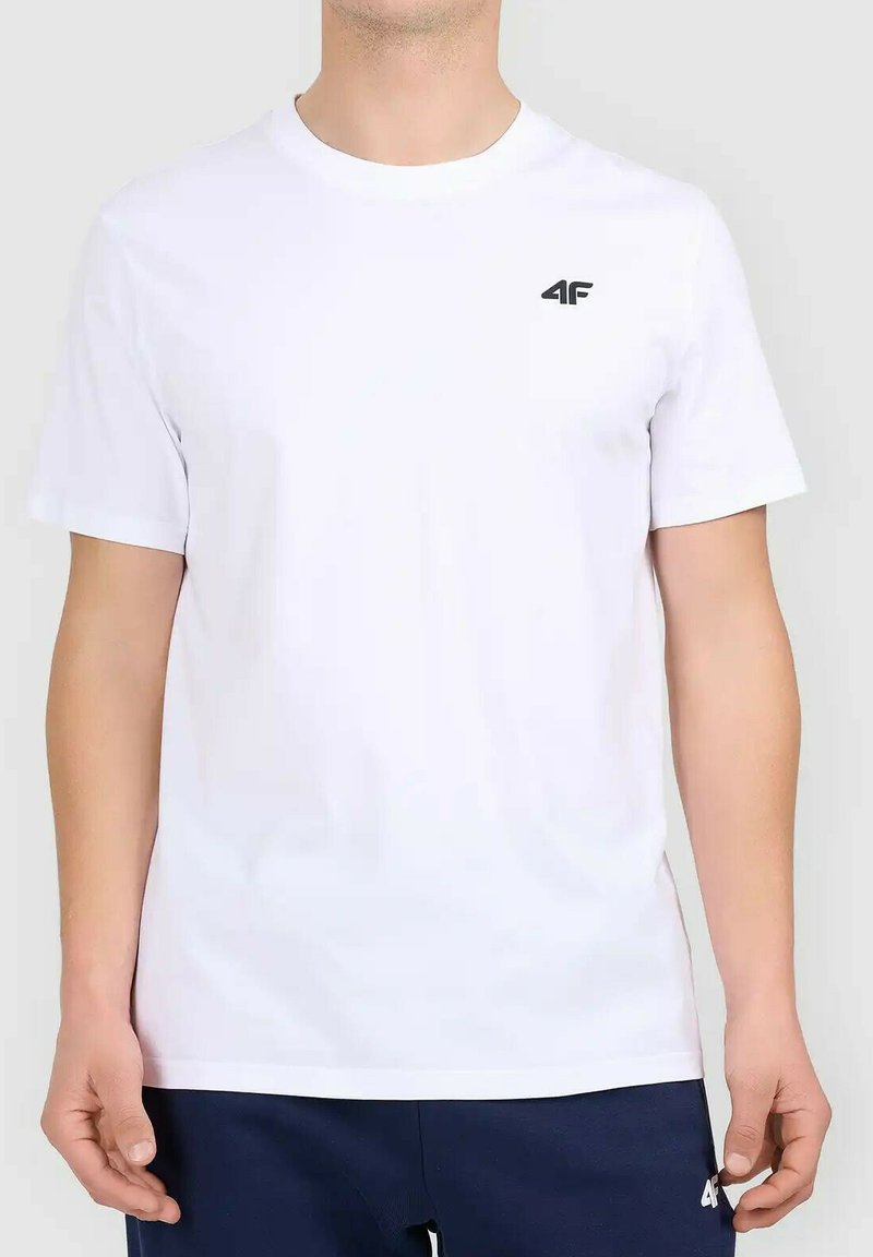Homme portant un t-shirt blanc uni à manches courtes avec un petit logo noir "4F" en haut à gauche, associé à un pantalon bleu foncé.