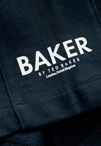 Sort stof med en glat tekstur, der pryder hvid branding: "BAKER BY TED BAKER" og "London, United Kingdom" i et rent, moderne skrifttype.