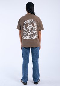 Camiseta de algodón marrón con un gráfico de signo de paz floral blanco en la parte posterior, combinada con jeans de mezclilla azul de corte recto.