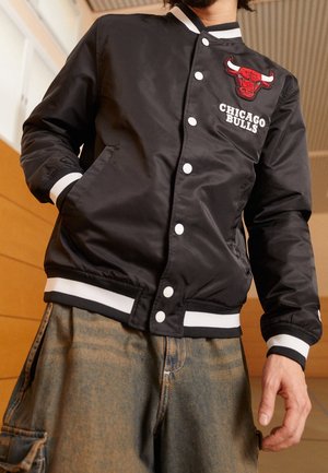 Personne portant une veste varsity noire des Chicago Bulls avec des boutons blancs et des poignets rayés, associée à un pantalon cargo marron ample.