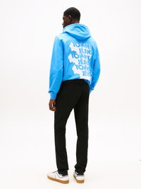 Blauwe hoodie met witte tekst "TOMMY JEANS" in een verspringend patroon op de achterkant, gecombineerd met zwarte broek en witte sneakers.