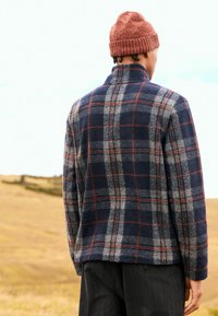 Marineblau karierte Fleecejacke mit grauen und roten Streifen, hohem Kragen und lässiger Passform. Getragen mit einer rostfarbenen Strickmütze. Strukturierten Stoff.