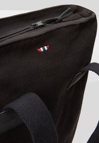 Sac cabas en tissu noir avec des poignées noires, une fermeture éclair et un petit détail cousu rouge, blanc et bleu près du bord supérieur.