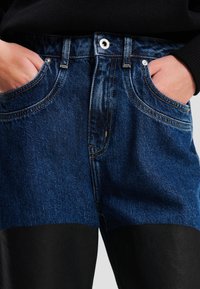 Jeanse iz jeansa z mešanico temno modre in črne tkanine. Značilnosti vključujejo standardni pas, pet žepov in vidne šive.