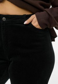 Pantaloni in velluto a coste neri con una sottile texture a coste, dotati di tasca laterale e chiusura con bottone metallico in vita.