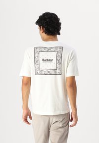 Bílá bavlněná trička s černobílým dekorativním potiskem na zádech, rámečkovaným do čtverce. Krátké rukávy, design s kulatým výstřihem.
