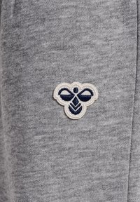 Grå bomullstyg med en texturerad yta, med en cirkulär broderad bi-logotyp i marinblått och vitt på övre vänstra sidan.