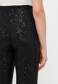 Czarne legginsy cekinowe o dopasowanym kroju, gładkiej strukturze i błyszczącej powierzchni. Widok z tyłu pokazuje szczegółowy wzór cekinów.