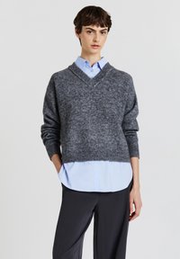 Grau gestrickter Pullover mit V-Ausschnitt, strukturiertem Stoff, überschnittenen Schultern und geripptem Saum. Trägt man über einem hellblauen Hemd mit Kragen und dunklen Hosen.