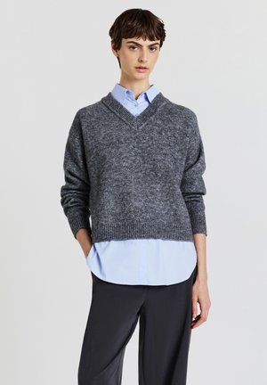 Pull en maille grise avec un col en V, tissu texturé, épaules tombantes et ourlet côtelé. Porté par-dessus une chemise à col bleu clair et un pantalon sombre.