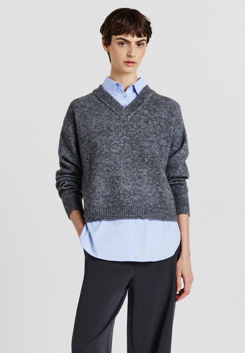 Grau gestrickter Pullover mit V-Ausschnitt, strukturiertem Stoff, überschnittenen Schultern und geripptem Saum. Trägt man über einem hellblauen Hemd mit Kragen und dunklen Hosen.