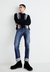 Polo negro de manga larga con rayas verticales grises, combinado con jeans de mezclilla azules y botas de ante negras, de pie contra un fondo blanco.