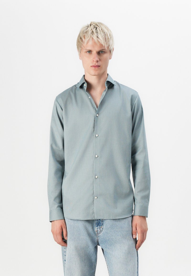 Camisa de botones azul claro con mangas largas, textura de tejido liso y cuello clásico, combinada con jeans de mezclilla claros.