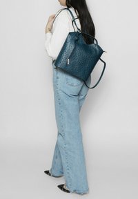 Zaino in pelle perforata blu navy con una forma strutturata, manici superiori e spallacci regolabili. Presenta una tasca frontale con zip.