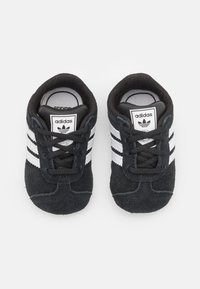 Černé semišové tenisky se třemi bílými pruhy, kulatou špičkou, šněrovacím designem a etiketou s logem na jazyku. Měkká textura vhodná na neformální nošení.