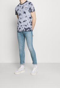 Miestenské tričko s farbením na modro a tmavo modré, krátke rukávy, okrúhly výstrih, s detailom žltého loga. Spárované s svetlomodrými úzkymi džínsami.