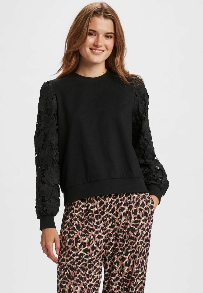 Felpa nera con maniche in pizzo testurizzato, scollatura rotonda e orlo a costine. Indossata con pantaloni beige e neri a stampa leopardata.
