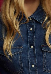 Camicia di denim con una tonalità blu scuro, caratterizzata da un colletto a punta, bottoni a pressione bianchi e due tasche sul petto con cuciture a contrasto.