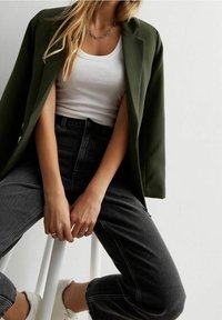 Olijfgroene blazer over een witte tank top, gecombineerd met hoog gesloten donkergrijze jeans en witte sneakers. Zilveren ketting zichtbaar.