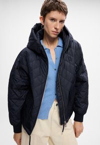 Gepufferte schwarze Steppjacke mit großer Kapuze, Reißverschluss und Seitentaschen. Kombiniert mit einem hellblauen gerippten Oberteil mit Knöpfen.