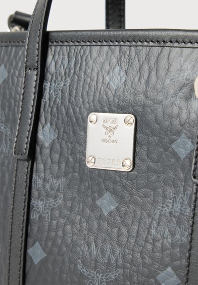 Μαύρη υφασμάτινη δερμάτινη τσάντα με επαναλαμβανόμενο μοτίβο λογότυπου MCM και ασημένια μεταλλική πλάκα που εμφανίζει το όνομα της μάρκας και τον σειριακό αριθμό.