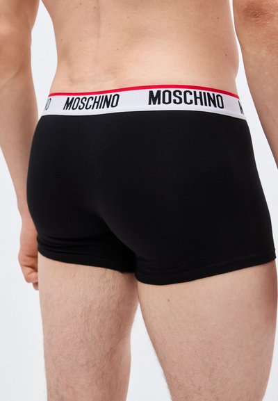 Άνδρας που φοράει μαύρα μποξεράκια με λευκή ελαστική ζώνη μέσης, επάνω στην οποία αναγράφεται το όνομα της μάρκας "Moschino" με μαύρα γράμματα και κόκκινο περίγραμμα στην κορυφή.