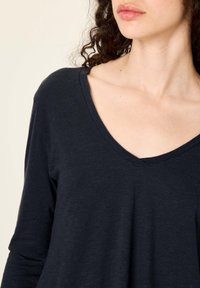 Moteris su garbanotais tamsiais plaukais, dėvinti juodą V formos iškirptės ilgomis rankovėmis marškinius, matoma nuo kaklo iki viršutinės liemens dalies, prie šviesaus fono.