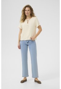 Blusa corta a maniche corte color crema con scollatura a chiave e fiocco, abbinata a jeans dritti azzurri e ballerine marroni. Texture liscia, design casual.