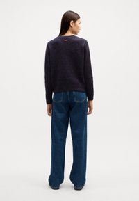 Marineblauer, gestrickter Pullover mit strukturiertem Finish, geripptem Saum und Rückenschild. Kombiniert mit hoch-taillierten, weit geschnittenen blauen Jeans.