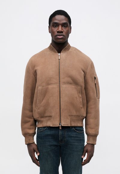 Férfi bézs nubuk bomber dzsekit visel, elülső cipzárral és cipzáras zsebbel az ujjon, sötétkék farmert párosítva, egy fehér háttér előtt áll.