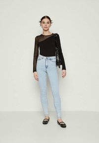 Crna majica dugih rukava s prozirnim lijevim ramenom, svijetloplave uske traperice i crne balerinke sa zakovicama; model pozira na jednostavnoj pozadini.