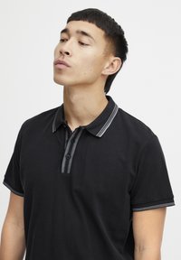 Svart polo skjorte med korte ermer, med en kontrastfarget stripet krage, en knappestolpe og et teksturert stoff.